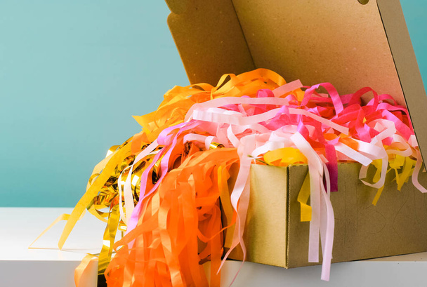 Paper garlands in a cardboard box against blue wall. Colorful holiday paper ribbons, sparkling paper, streamers. Celebration curly mixed golden, yellow, pink tapes. Congratulation, festive event, fun. - Photo, Image