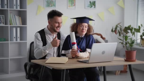 distance learning, cheerful young couple in academic clothes celebrating graduation ceremony and diploma by video link on laptop - Video