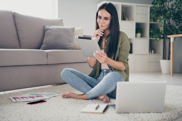 Full size photo of attractive positive lady sit on floor barefoot hand on chin look minded tablet working home indoors - Photo, image