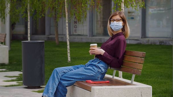 Business Caucasian Woman wearing medical mask takes a break. Outdoor Relaxing break from work enjoys sunny spring summer day coffee break. Lady looks thoughtful and enjoying coffee.  - Foto, Imagem