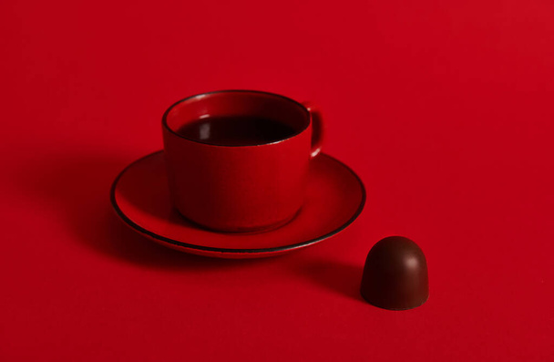 Closeup of a ceramic cup of coffee on a saucer and a chocolate praline on red background with copy space . Concept of chocolate day - Photo, Image