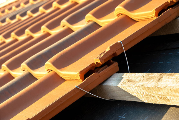 Detailní záběr kovové montážní kotvy pro montáž žlutých keramických střešních tašek namontovaných na dřevěných deskách kryjících bytovou střechu budovy ve výstavbě. - Fotografie, Obrázek