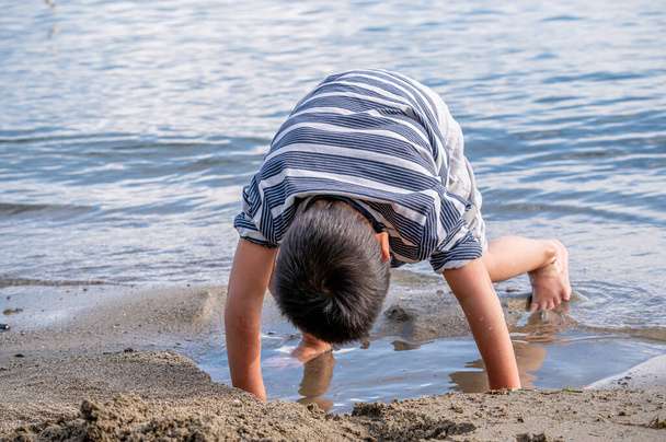 Lapsi leikkii hiekkaa rannalla. Pieni aasialainen poika rennoissa vaatteissa veden äärellä Sveitsissä. Hyvää lapsuutta.. - Valokuva, kuva