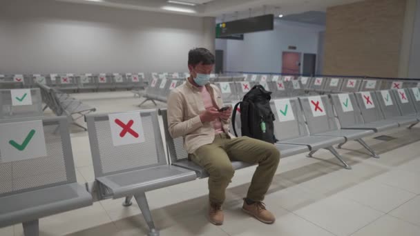 キプロス、パフォス市の空港で空のターミナルに座って、キャンセル便で動揺保護マスクを身に着けている男は、彼の家族にメッセージを書き込み、 Covid 19流行のコロナウイルスパンデミックのために - 映像、動画