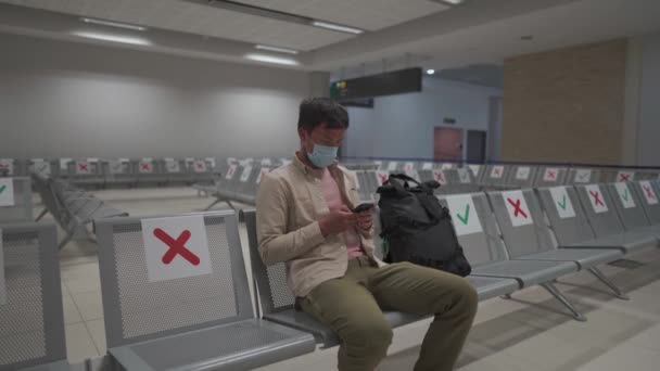 Путешественник сидит в международном аэропорту Кипра в Пафосе во время кризиса COVID-19, сидит в терминале на сиденье с социальной разметкой расстояния в маске. Использование телефона в ожидании рейса авиакомпании - Кадры, видео
