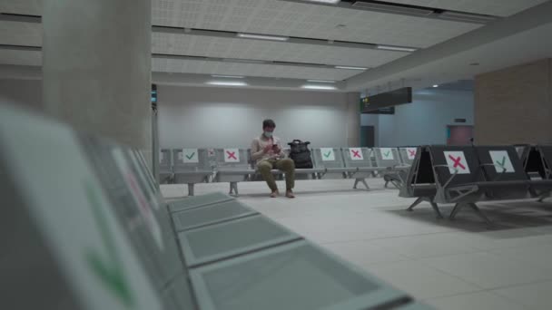 旅行者は、 COVID-19危機の間にパフォスのキプロスの国際空港に座って、マスクを身に着けている社会的距離マーキングと座席のターミナルに座っています。飛行機に乗るのを待っている間に電話を使う - 映像、動画