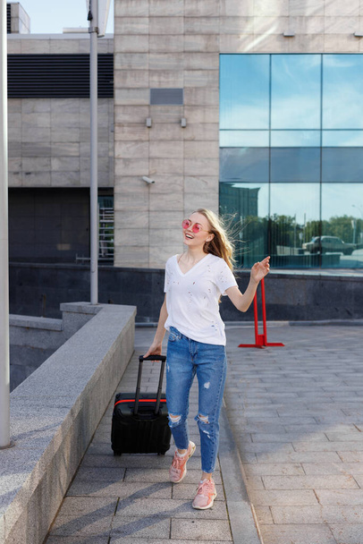 空港ターミナルの近くで笑顔のスーツケースとピンクのメガネの魅力的なブロンド。旅行2021 - 写真・画像