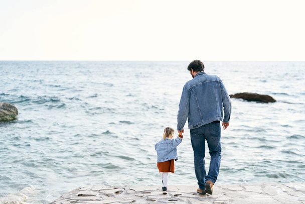 Papá con una chaqueta de mezclilla y jeans lleva a una niña por la mano a lo largo de la costa rocosa hasta el agua. Vista trasera - Foto, Imagen