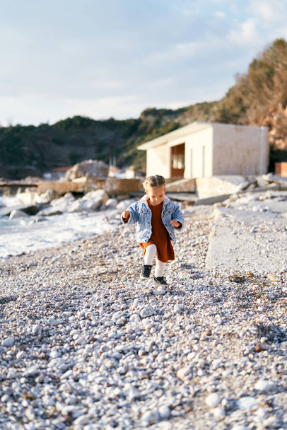 Dziewczynka w sukience i dżinsowej kurtce spaceruje po żwirowej plaży patrząc na jej stopy na tle morza, skał i budynku. - Zdjęcie, obraz