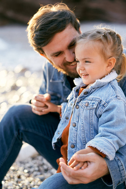 Papá sonriente se sienta agachándose ante una niña riendo en una playa de guijarros. Primer plano - Foto, Imagen