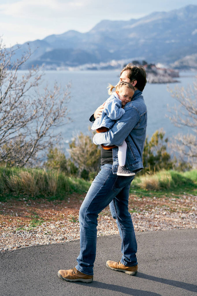 Papà abbraccia una bambina sorridente al petto, camminando lungo la strada sullo sfondo del mare, delle montagne e dell'isola di Sveti Stefan - Foto, immagini