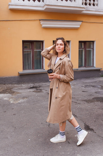 Young smiling millennial woman with wild hair dressed in an autumn coat walking with a cup of coffee to go near the old building. - Фото, зображення