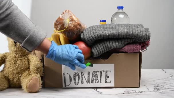 коричневая картонная коробка с едой, одеждой и игрушками на столе, рука в голубой перчатке прикрепляет лист бумаги с надписью пожертвовать. Помощь бедным, нуждающимся - Кадры, видео