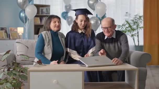 family with graduate celebrating end of school year online and preparing for graduation ceremony via video link with teacher sitting in living room with balloons - Video