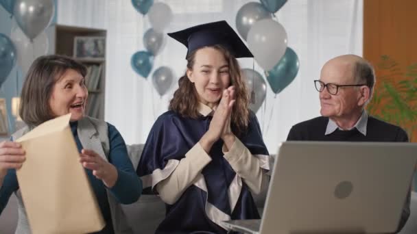 graduate in hat and gown together with parents celebrate graduation from university during ceremony by video link on laptop sitting at home background of balloons - Video