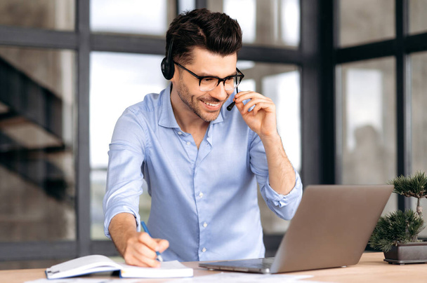 A friendly caucasian man with glasses, office worker or consultant, stands near a workplace, uses a headset and laptop for online consultation or negotiations, communicates via video communication - Photo, Image