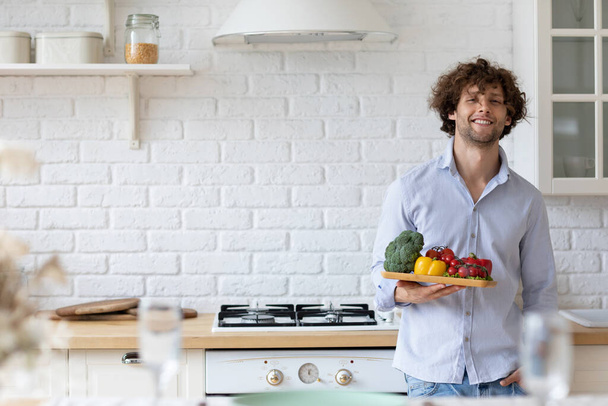 自宅のキッチンに立ちながらカメラを見て笑顔でカジュアルな服装で野菜を持つハンサムな男。彼の健康の世話を. - 写真・画像