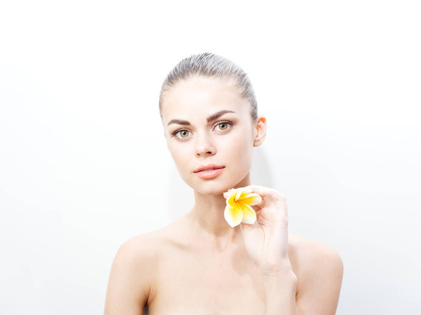 mujer bonita con una flor amarilla cerca de la cara sobre un fondo claro hombros desnudos - Foto, Imagen