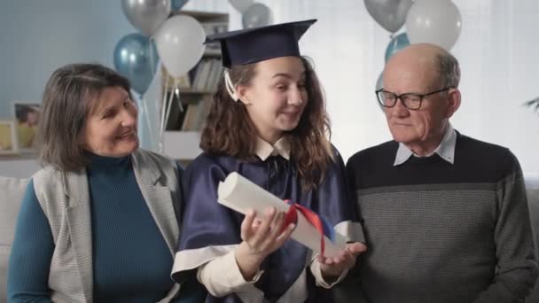 retrato familia feliz con hija graduada en una gorra académica y vestido con diploma en las manos se regocija al final del año escolar - Imágenes, Vídeo