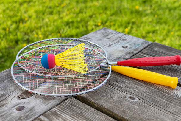 Badminton game rackets and shuttlecock on wooden table with green grass backgroud in the park on a sunny summer day. Active lifestyle concept. Fun outdoors leisure activities for a family. - Photo, Image