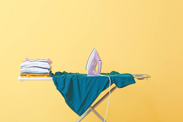 Board with electric iron and clean clothes on color background - Foto, Imagen