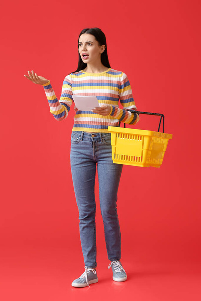 Unzufriedene junge Frau mit Warenkorb und Produktliste auf farbigem Hintergrund - Foto, Bild