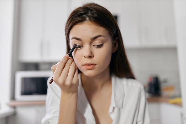 woman makes makeup in the kitchen indoors with eyeliner in hand - Photo, Image