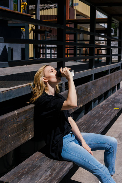 A young beautiful woman is sitting on a bench and drinking coffee. The girl is sitting outside resting in the bright sun. Walking around the city after the pandemic. - Photo, Image