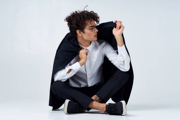 curly-haired guy straightens his jacket over his shoulders and sits on the floor in sneakers and in a suit - Photo, Image