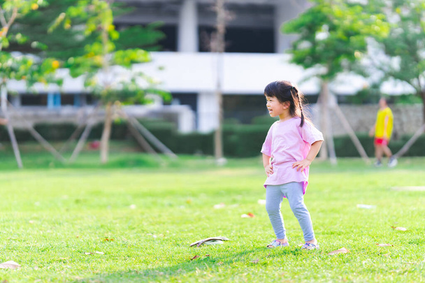 A 4 year old Asian girl enjoys exercising. Bright green lawn. Public areas for sporting activities. Happy child handle waist and spins. Sweet smile and laughing. Funny kid wear pink T-shirt. - Foto, afbeelding