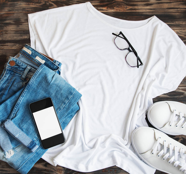 Stylish female clothes set. Woman outfit on wooden background. White t-shirt, white sneakers and sunglasses. Flat lay, top view - 写真・画像