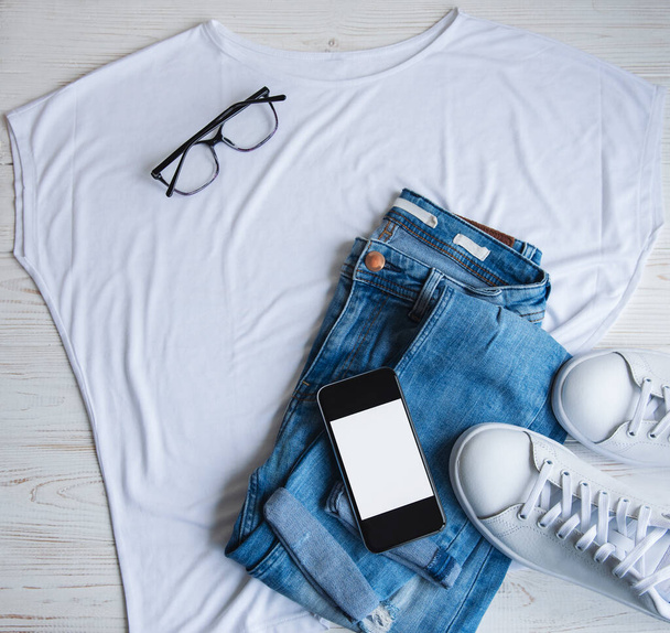 Stylish female clothes set. Woman outfit on  wooden background. Blue jeans, white t-shirt, white sneakers and sunglasses. Flat lay, top view - 写真・画像