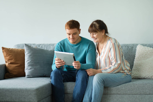 son teaching his mother to use tablet. older people using technology. Cheerful elderly woman sitting on the sofa next to his adult son - Photo, Image