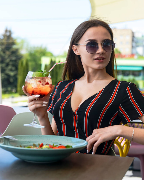 Young attractive woman eating mixed caprese salad with strawberries and drinking aperol spritz soda cocktail in outdoor restaurant. Eat out concept, eating alone due to worldwide pandemic. - 写真・画像