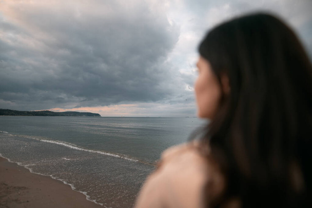 Μοναχική γυναίκα περπατά στην παραλία. Ντυμένη μελαχρινή που περπατάει δίπλα στη θάλασσα το βράδυ. Ένα κορίτσι τις ανοιξιάτικες διακοπές δίπλα στη θάλασσα. - Φωτογραφία, εικόνα