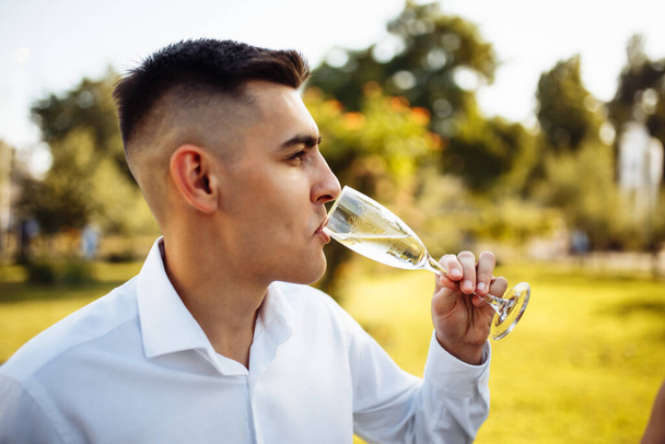 A young man drinks champagne at a luxury wedding. A man with a glass in his hands at the celebration. Summer portrait of a man in a festive mood. Celebrating a wedding, anniversary, birthday - Fotografie, Obrázek