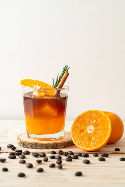 A glass of iced americano black coffee and layer of orange and lemon juice decorated with rosemary and cinnamon on wood background - Фото, зображення