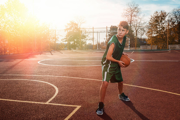 Sports and basketball. A young teenager in a black tracksuit poses with a basketball in his hands and is ready to throw. In the background, a basketball court. Copy space. Sunlight. - Photo, image