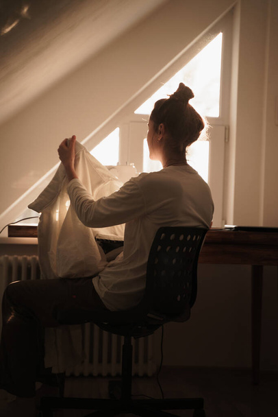 A shallow focus of a woman sitting by a bright window while fixing the white curtains - Foto, afbeelding
