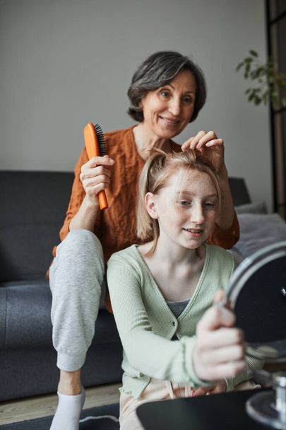 Жінка посміхається і чистить волосся для своєї веснянки онуки у вітальні
 - Фото, зображення