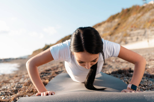 A sport fogalma. Egy fiatal barna nő fekvőtámaszt csinál egy sportszőnyegen. A háttérben hegyek, tenger és part. - Fotó, kép