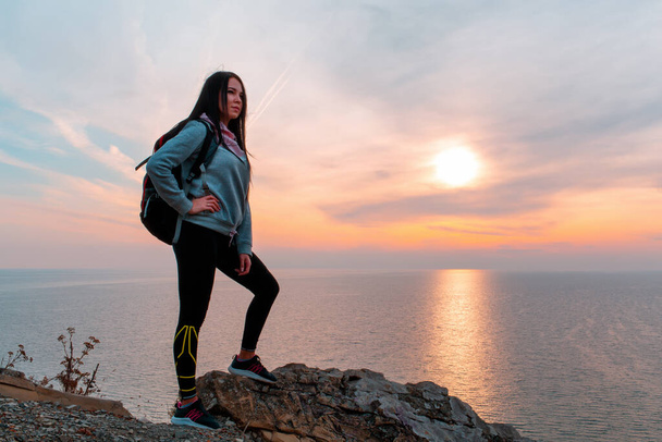 The concept of Hiking and sports recreation. A woman with a backpack on her back poses against the sea and sky. Sunset. Copy space. - Foto, imagen