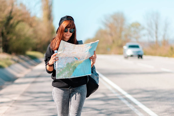 Hitchhiking trips. Portrait of young smiling woman in a cap and sunglasses, holding a paper map. In the background, a road with driving cars is blurred. The concept of local travel. - Foto, Bild