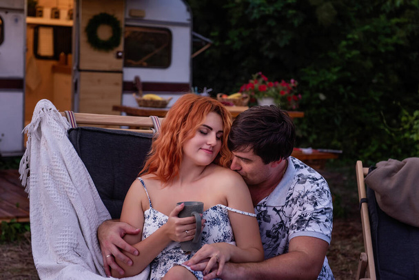 Middle-aged man kisses the shoulder of young red-haired woman. Loving couple resting in the evening, hugging behind the countryside at the motor house. Budget Travel on Trailer Track for the weekend - Photo, image