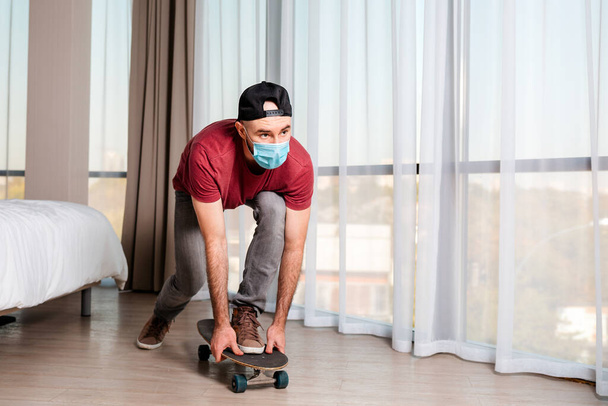 A young man in a medical mask, riding a longboard. Home interior. Indoors. Concept of quarantine and activity. - Foto, immagini