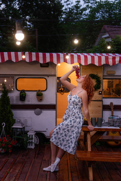 Young red-haired woman in white, flower sundress eats ripe grapes near the motor at home. Trailer Track is decorated with garlands of lamps, petunias blooming. Happy girl smiles, enjoys the trip - Foto, immagini