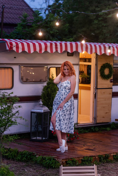 Young red-haired woman in white, floral sundress is resting by the motor at home in the evening. Trailer Track is decorated with garlands of lamps, blooming petunias. Happy girl smiles enjoys the trip - Φωτογραφία, εικόνα