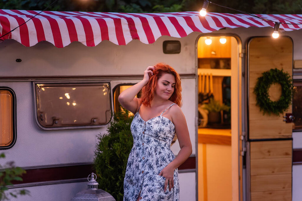 Young red-haired woman in white, floral sundress is resting by the motor at home in the evening. Trailer Track is decorated with garlands of lamps, blooming petunias. Happy girl smiles enjoys the trip - Foto, afbeelding