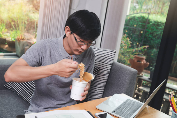 Asian man eating instant noodles while working on laptop at his home office. - Foto, Imagen