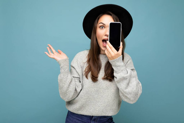 positive lady student wearing black hat and grey sweater holding mobilephone showing smartphone isolated on background looking at camera with hand - Fotografie, Obrázek